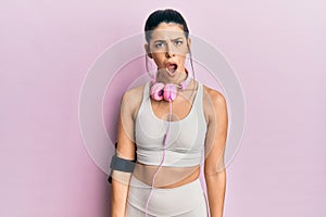 Young hispanic woman wearing gym clothes and using headphones in shock face, looking skeptical and sarcastic, surprised with open