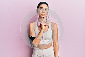 Young hispanic woman wearing gym clothes and using headphones thinking concentrated about doubt with finger on chin and looking up