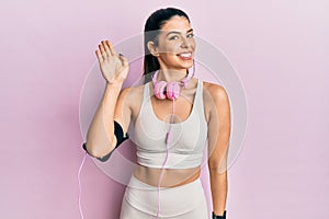 Young hispanic woman wearing gym clothes and using headphones waiving saying hello happy and smiling, friendly welcome gesture