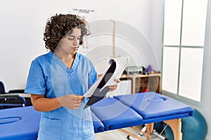 Young hispanic woman wearing physio therapist uniform reading paperwork at clinic