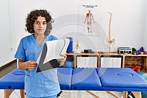 Young hispanic woman wearing physio therapist uniform reading paperwork at clinic