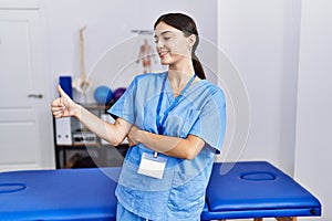 Young hispanic woman wearing physiotherapist uniform standing at clinic looking proud, smiling doing thumbs up gesture to the side