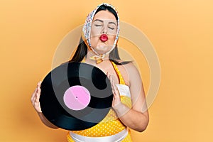 Young hispanic woman wearing pin up style holding vinyl disc looking at the camera blowing a kiss being lovely and sexy