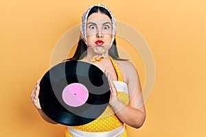 Young hispanic woman wearing pin up style holding vinyl disc puffing cheeks with funny face