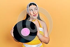 Young hispanic woman wearing pin up style holding vinyl disc smiling looking to the side and staring away thinking