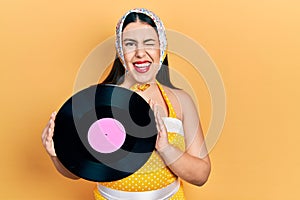 Young hispanic woman wearing pin up style holding vinyl disc winking looking at the camera with sexy expression, cheerful and