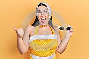 Young hispanic woman wearing pin up style pointing thumb up to the side smiling happy with open mouth