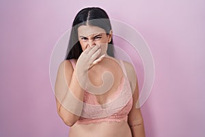 Young hispanic woman wearing pink bra smelling something stinky and disgusting, intolerable smell, holding breath with fingers on