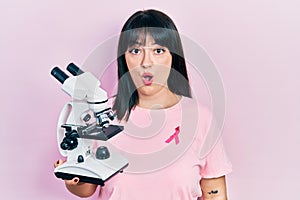Young hispanic woman wearing pink cancer ribbon on shirt holding microscope scared and amazed with open mouth for surprise,
