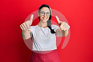 Young hispanic woman wearing professional waitress apron approving doing positive gesture with hand, thumbs up smiling and happy