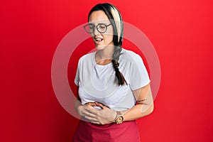 Young hispanic woman wearing professional waitress apron with hand on stomach because nausea, painful disease feeling unwell