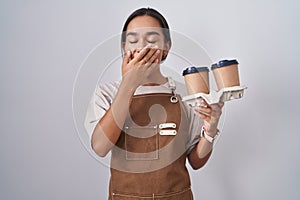 Young hispanic woman wearing professional waitress apron holding coffee bored yawning tired covering mouth with hand