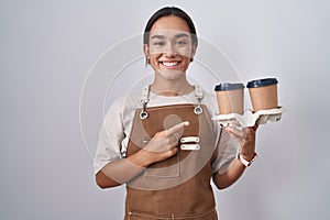 Young hispanic woman wearing professional waitress apron holding coffee cheerful with a smile on face pointing with hand and