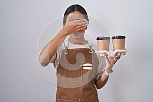 Young hispanic woman wearing professional waitress apron holding coffee covering eyes with hand, looking serious and sad