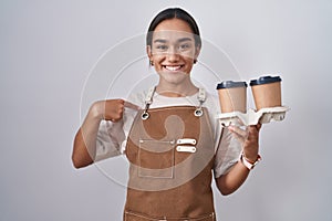 Young hispanic woman wearing professional waitress apron holding coffee looking confident with smile on face, pointing oneself