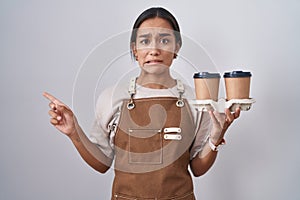 Young hispanic woman wearing professional waitress apron holding coffee pointing aside worried and nervous with forefinger,