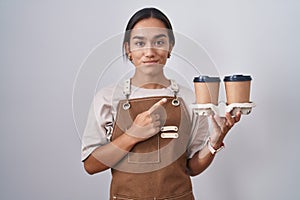 Young hispanic woman wearing professional waitress apron holding coffee pointing with hand finger to the side showing