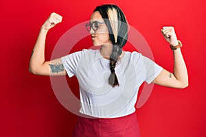 Young hispanic woman wearing professional waitress apron showing arms muscles smiling proud