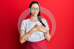 Young hispanic woman wearing professional waitress apron smiling with hands on chest with closed eyes and grateful gesture on face