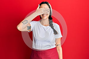 Young hispanic woman wearing professional waitress apron smiling and laughing with hand on face covering eyes for surprise