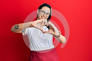 Young hispanic woman wearing professional waitress apron smiling in love doing heart symbol shape with hands