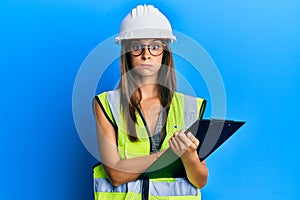 Young hispanic woman wearing safety helmet holding clipboard puffing cheeks with funny face