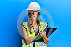 Young hispanic woman wearing safety helmet holding clipboard in shock face, looking skeptical and sarcastic, surprised with open