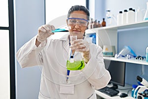 Young hispanic woman wearing scientist uniform pouring liquid on test tube at laboratory