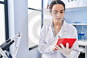 Young hispanic woman wearing scientist uniform using touchpad at laboratory