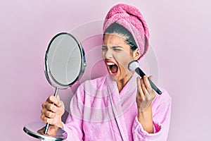 Young hispanic woman wearing shower bathrobe using makeup brush angry and mad screaming frustrated and furious, shouting with