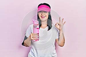Young hispanic woman wearing sportswear drinking bottle of water doing ok sign with fingers, smiling friendly gesturing excellent