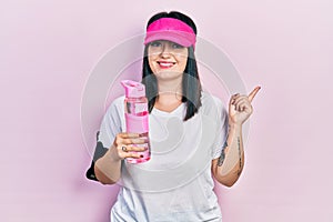 Young hispanic woman wearing sportswear drinking bottle of water smiling happy pointing with hand and finger to the side