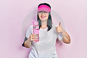 Young hispanic woman wearing sportswear drinking bottle of water smiling happy and positive, thumb up doing excellent and approval