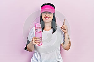 Young hispanic woman wearing sportswear drinking bottle of water smiling with an idea or question pointing finger with happy face,