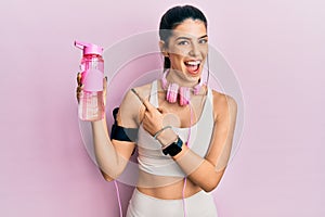 Young hispanic woman wearing sportswear, drinking bottle of water and using headphones smiling happy pointing with hand and finger