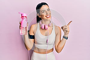 Young hispanic woman wearing sportswear, drinking bottle of water and using headphones smiling happy pointing with hand and finger