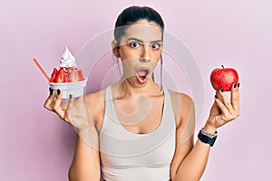 Young hispanic woman wearing sportswear holding apple and ice cream afraid and shocked with surprise and amazed expression, fear