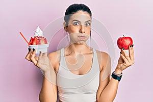 Young hispanic woman wearing sportswear holding apple and ice cream puffing cheeks with funny face