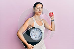 Young hispanic woman wearing sportswear holding weighing machine and apple looking at the camera blowing a kiss being lovely and