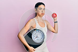 Young hispanic woman wearing sportswear holding weighing machine and apple puffing cheeks with funny face