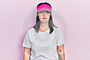 Young hispanic woman wearing sportswear and sun visor cap with serious expression on face