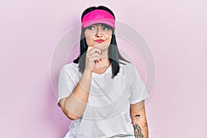 Young hispanic woman wearing sportswear and sun visor cap serious face thinking about question with hand on chin, thoughtful about