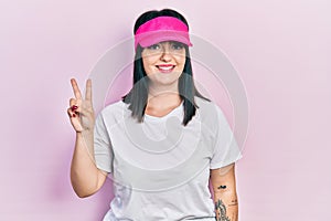 Young hispanic woman wearing sportswear and sun visor cap smiling with happy face winking at the camera doing victory sign