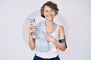Young hispanic woman wearing sportswear and towel drinking bottle of water smiling happy pointing with hand and finger