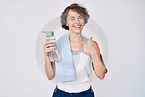 Young hispanic woman wearing sportswear and towel drinking bottle of water smiling happy and positive, thumb up doing excellent