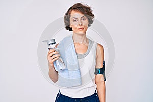 Young hispanic woman wearing sportswear and towel drinking bottle of water thinking attitude and sober expression looking self
