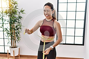 Young hispanic woman wearing sportswear using tape measure at sport center winking looking at the camera with sexy expression,