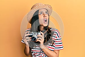 Young hispanic woman wearing summer hat holding vintage camera angry and mad screaming frustrated and furious, shouting with anger