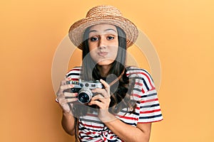 Young hispanic woman wearing summer hat holding vintage camera puffing cheeks with funny face