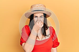 Young hispanic woman wearing summer hat smelling something stinky and disgusting, intolerable smell, holding breath with fingers